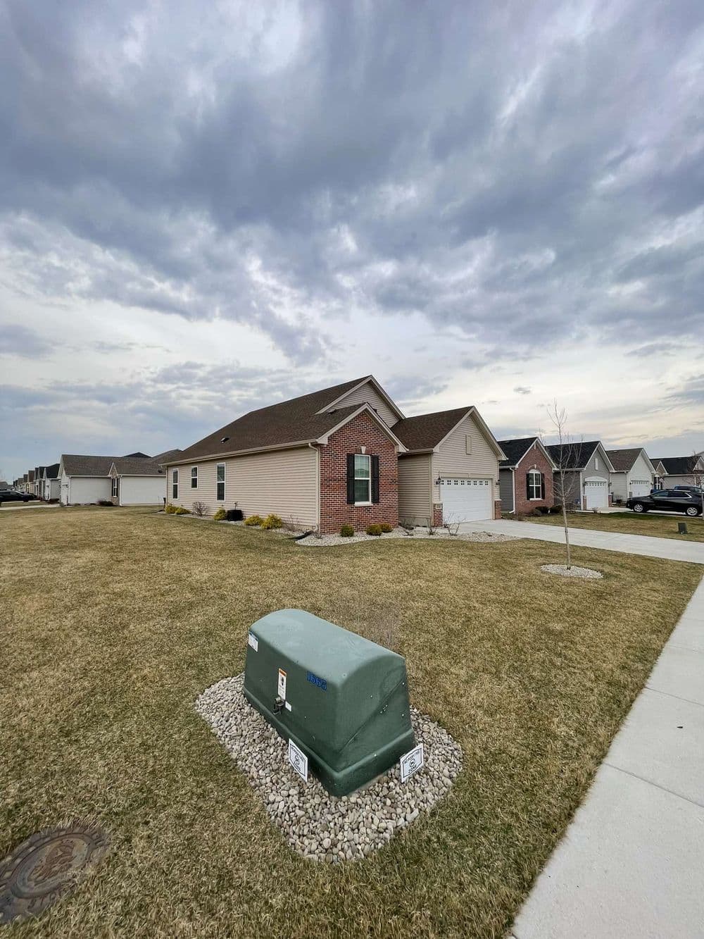 Single-story brick house with green utility box in yard under cloudy sky. Residential neighborhood.