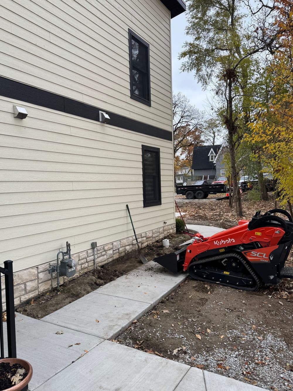 Small red excavator parked beside a house, near a freshly dug trench and concrete walkway.