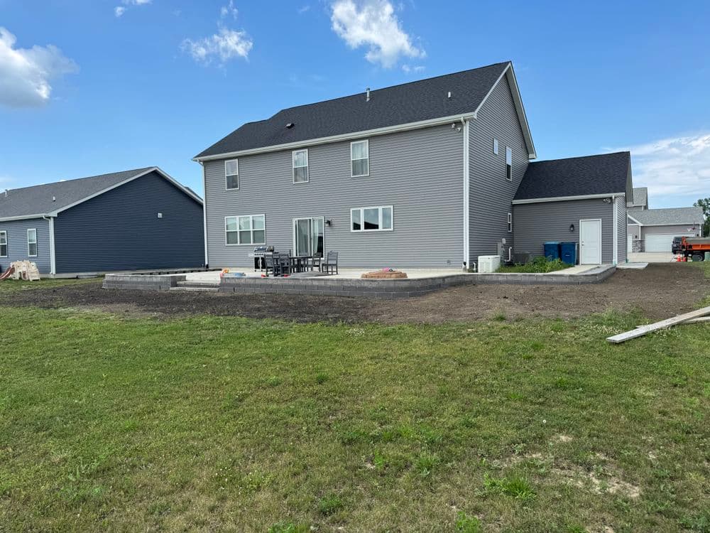 Two-story gray house with patio and green lawn under a blue sky. New construction site nearby.