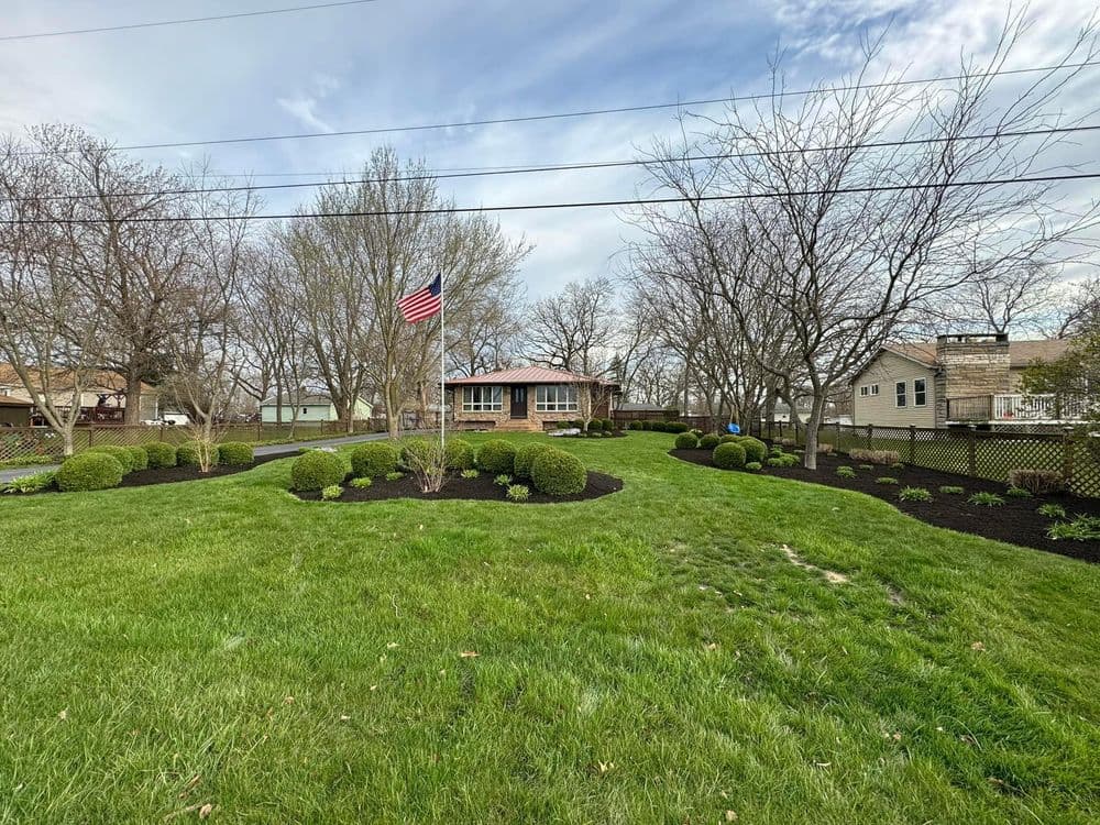 Lush green yard with neatly trimmed bushes, flagpole, and residential homes in the background.