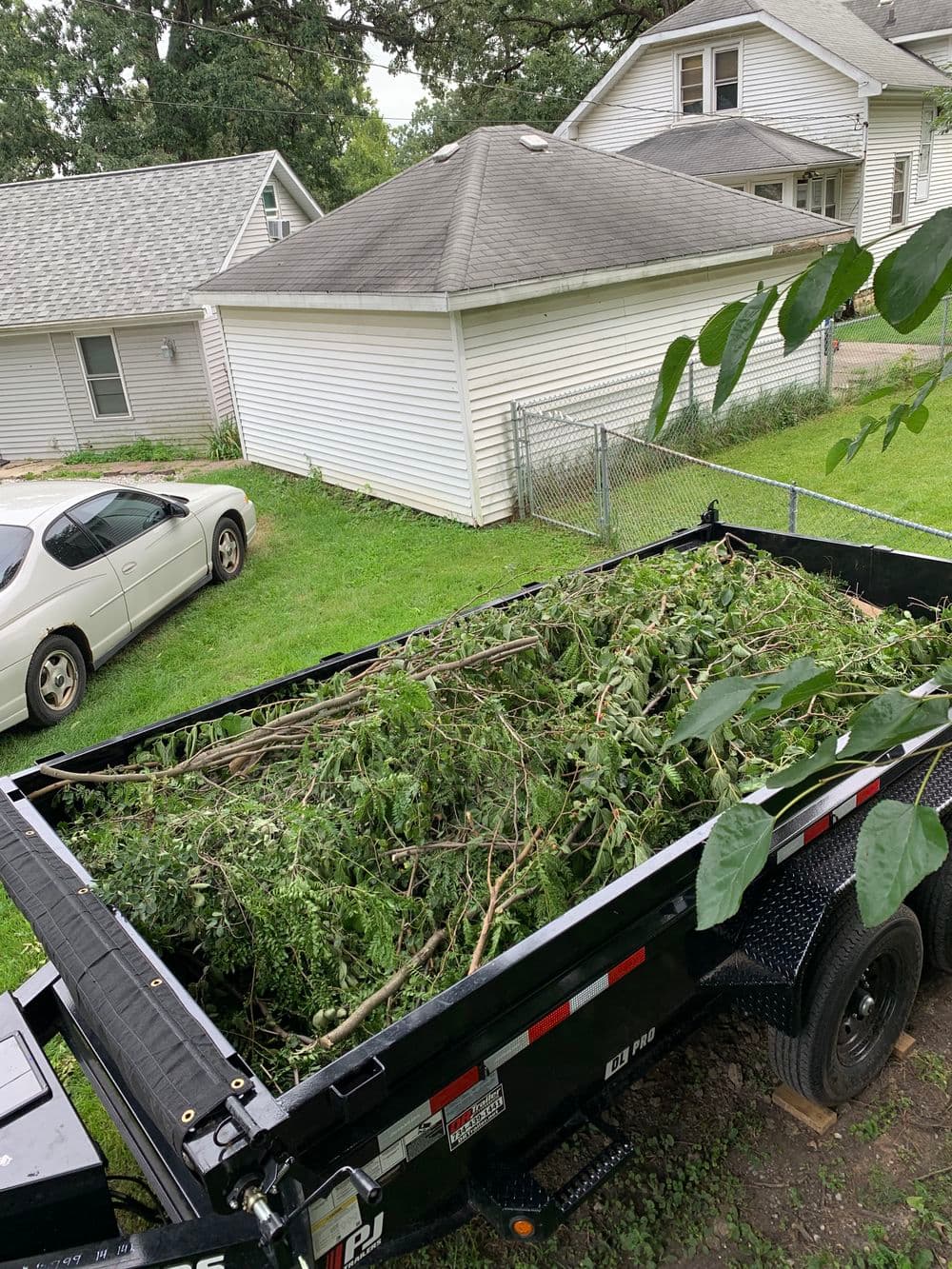 Dump trailer filled with tree branches parked in residential yard. Nearby houses visible.