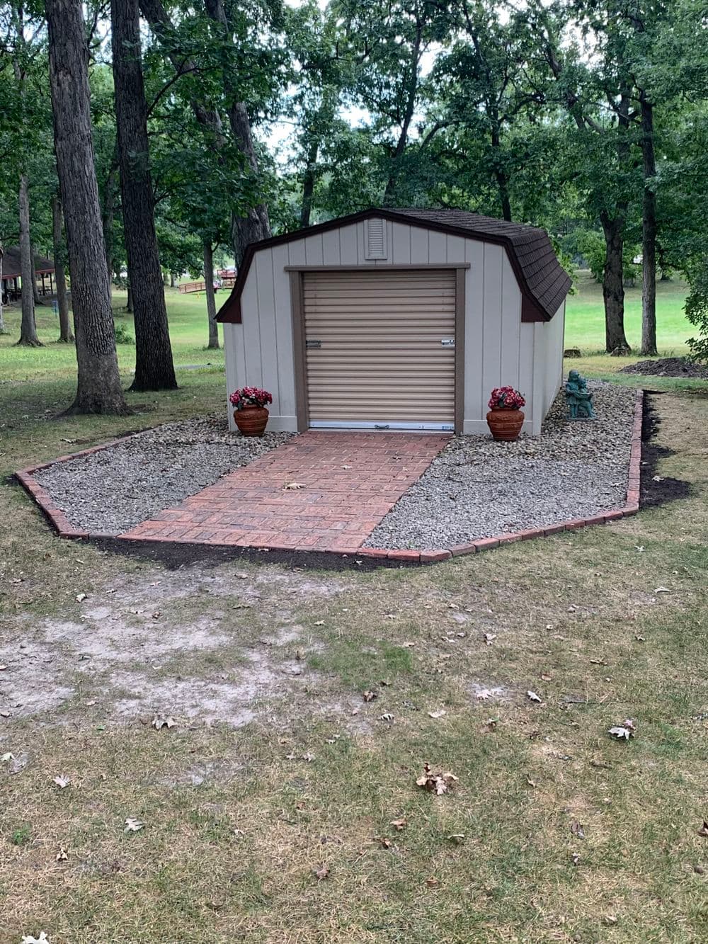 Garden shed with a gravel path and potted plants surrounded by grass and trees.