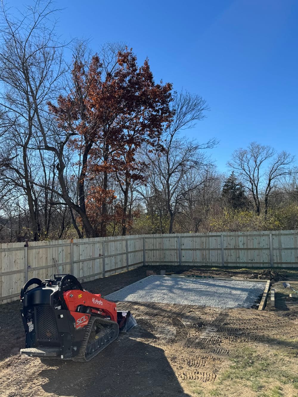 Red compact track loader on gravel area with wooden fence and trees under clear blue sky.
