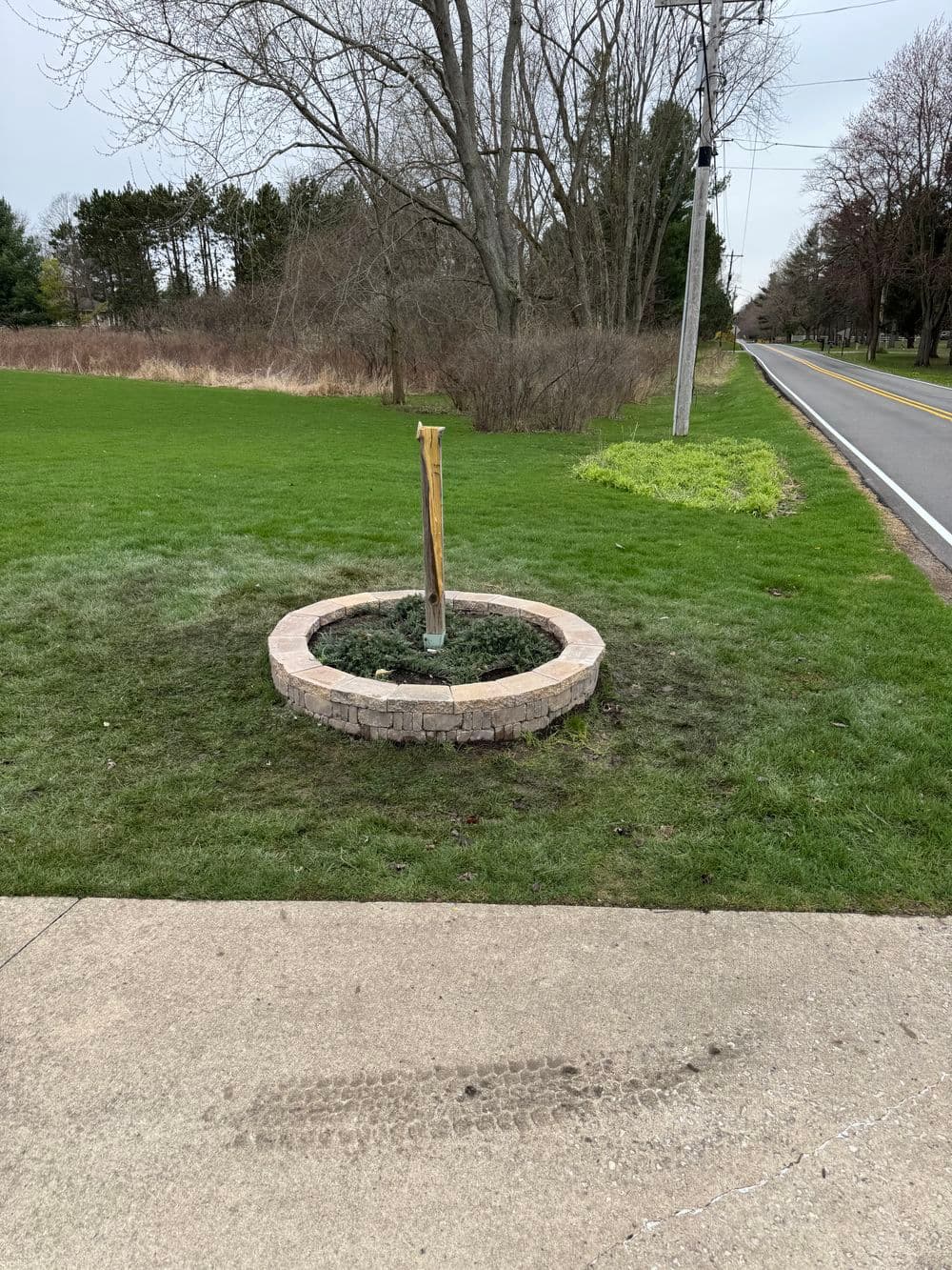 Wooden post in a circular stone planter by a grassy area near a road on a cloudy day.