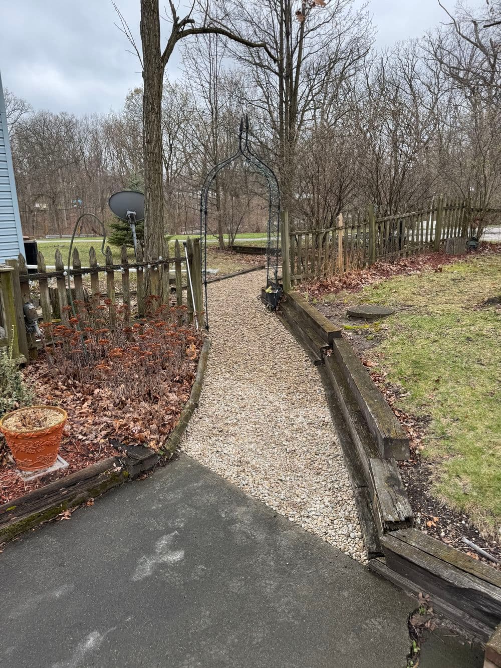 Gravel path leading to a garden archway surrounded by trees and a wooden fence.