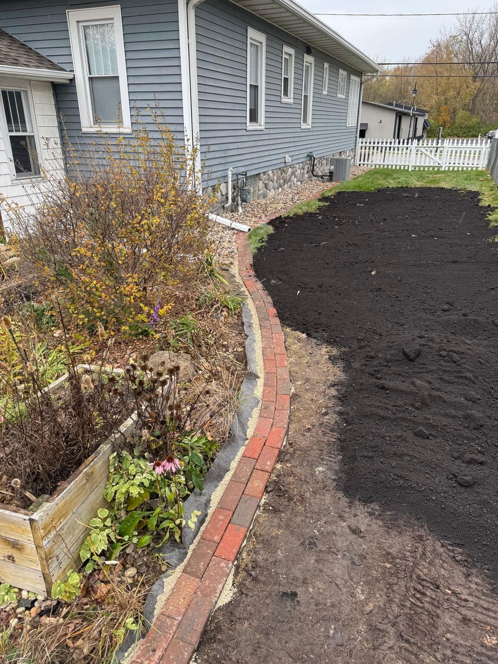 Garden area with newly prepared soil, bordered by a brick walkway and landscaped plants.