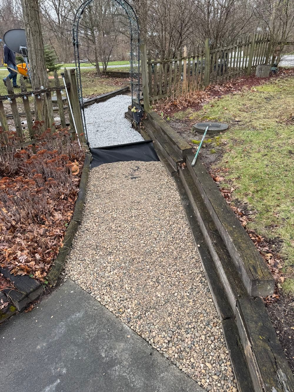 Gravel walkway leading to an archway in a garden with bare trees and wooden fence.