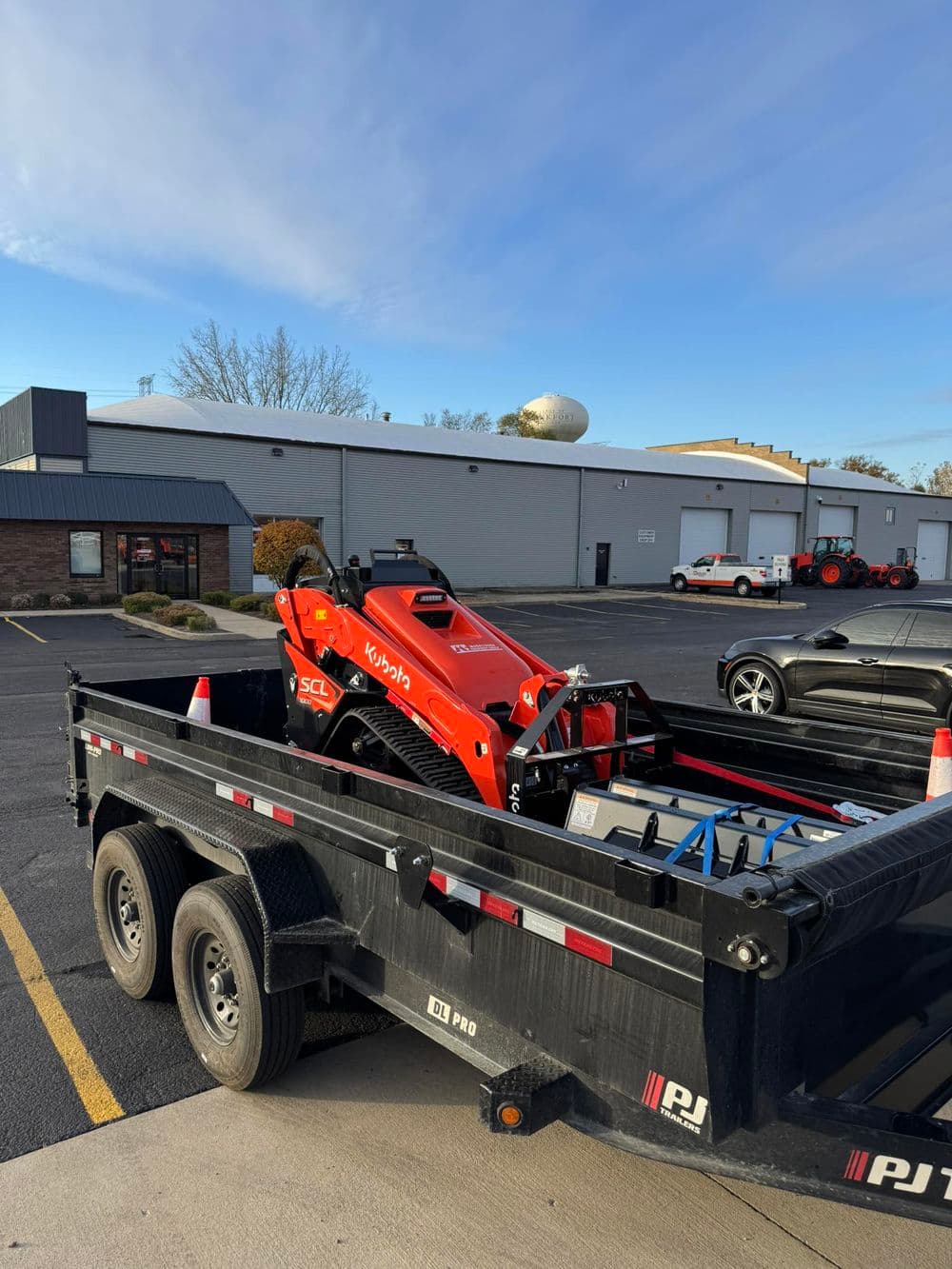 Compact tractor loaded on a black trailer in front of a commercial building.